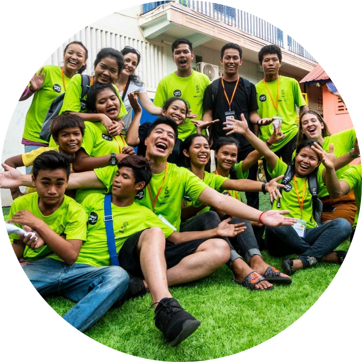Group of people wearing matching green shirts sitting together on grass outdoors, posing in front of a building during a team or group activity.
