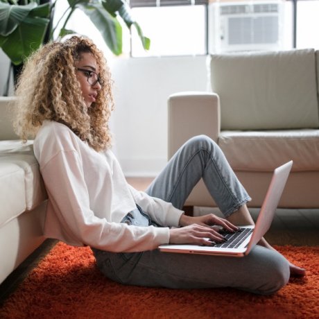 Person working on a laptop while sitting on a living room floor, leaning against a sofa in a bright, modern home with natural light and indoor plants.