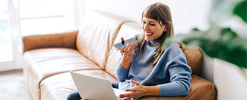 A woman smiling while talking on a smartphone and using a laptop on a tan leather couch.