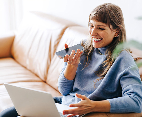 A woman smiling while talking on a smartphone and using a laptop on a tan leather couch.