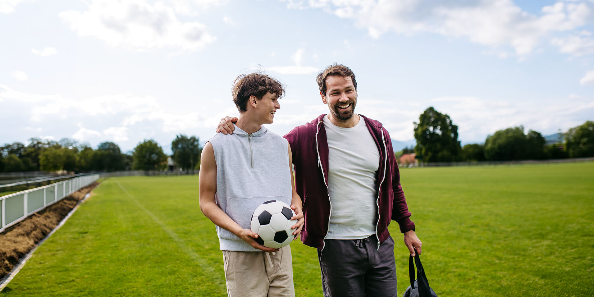 Father walking with his teenage son on a soccer field, with his arm around his shoulder while the son carries a soccer ball.