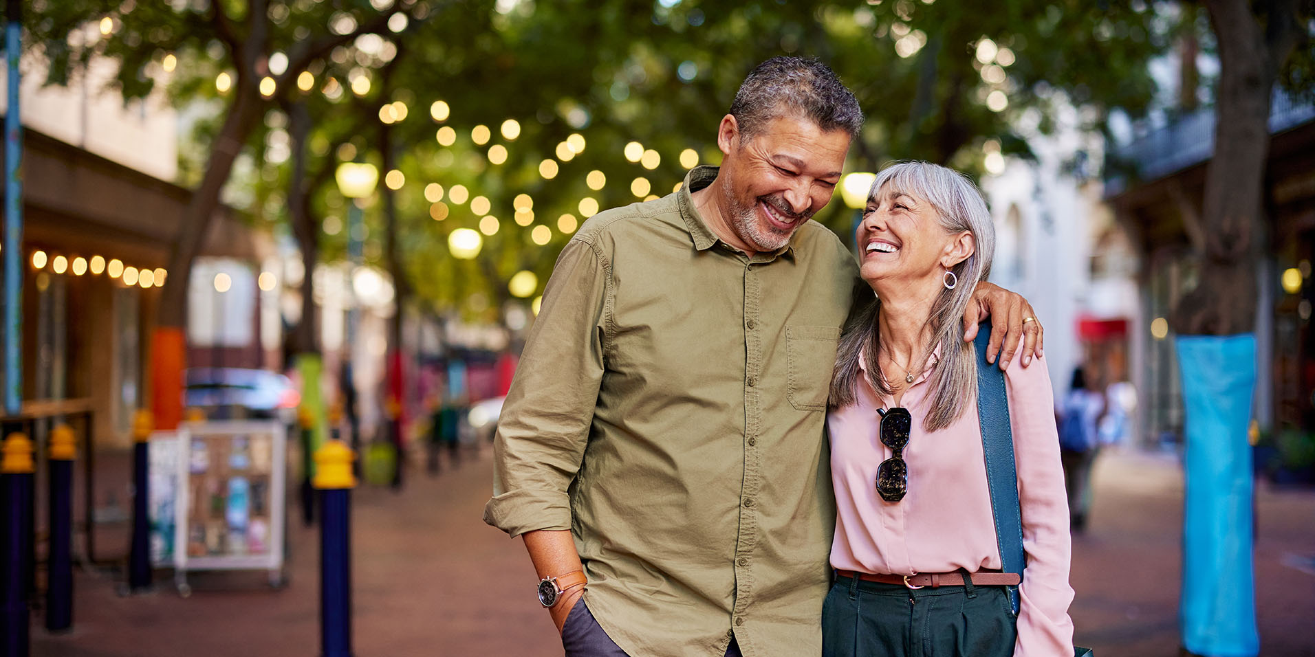 A middle-aged couple smiling and walking together along an outdoor shopping street lined with fairy lights.