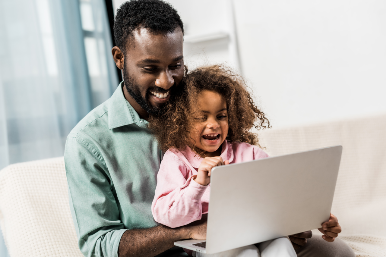 smiling african american child sitting with father on couch and using laptop 